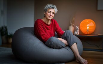 A woman with short, curly gray hair sits comfortably on a large gray bean bag, wearing a red sweater and light pants. A warm orange lamp and small decorations are on a table in the softly lit background.