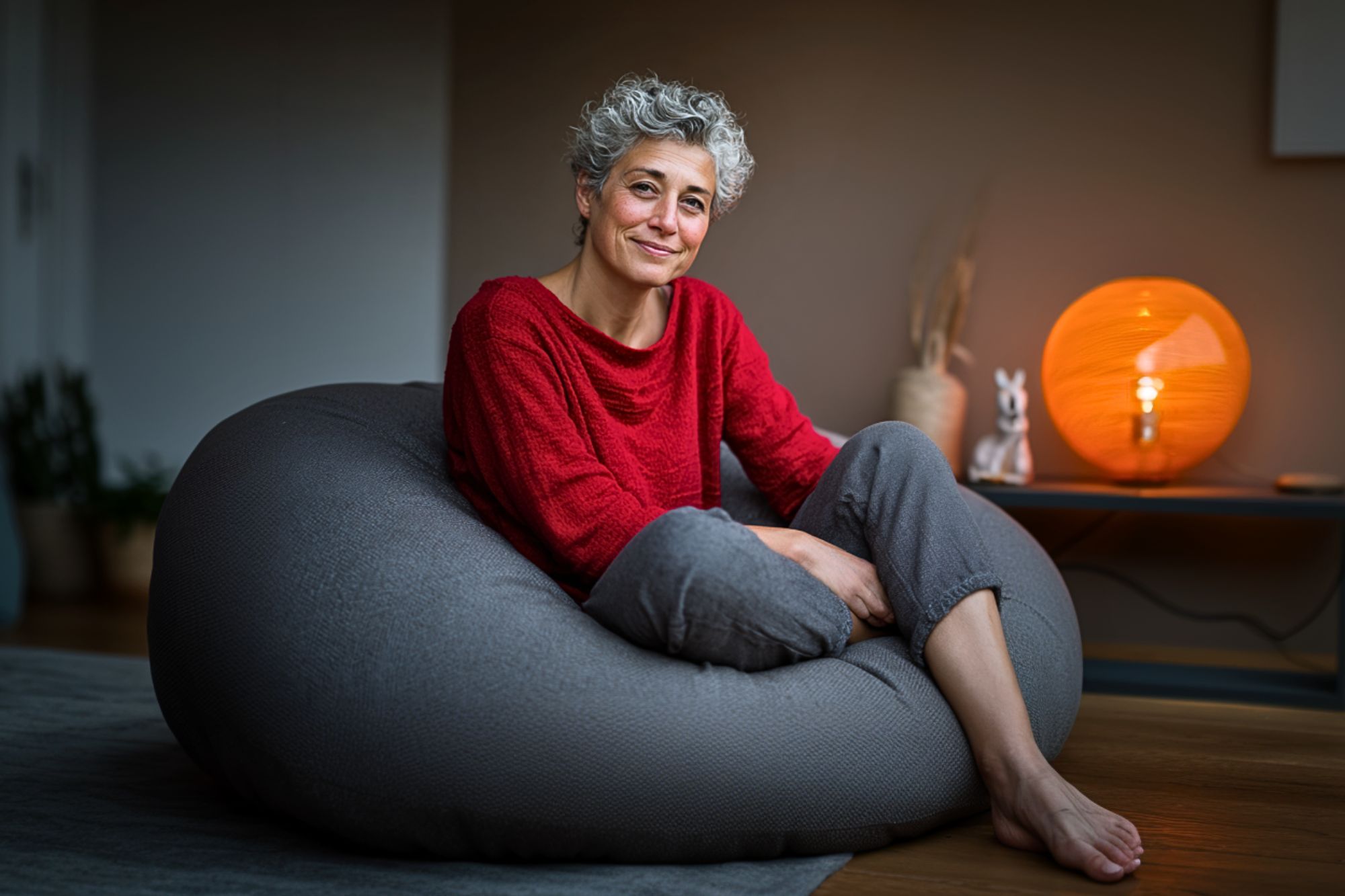 A woman with short, curly gray hair sits comfortably on a large gray bean bag, wearing a red sweater and light pants. A warm orange lamp and small decorations are on a table in the softly lit background.