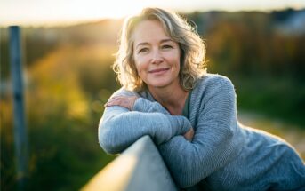 A woman with wavy gray hair and a gray sweater leans on a wooden fence, smiling gently outdoors at sunset with a blurred natural background.