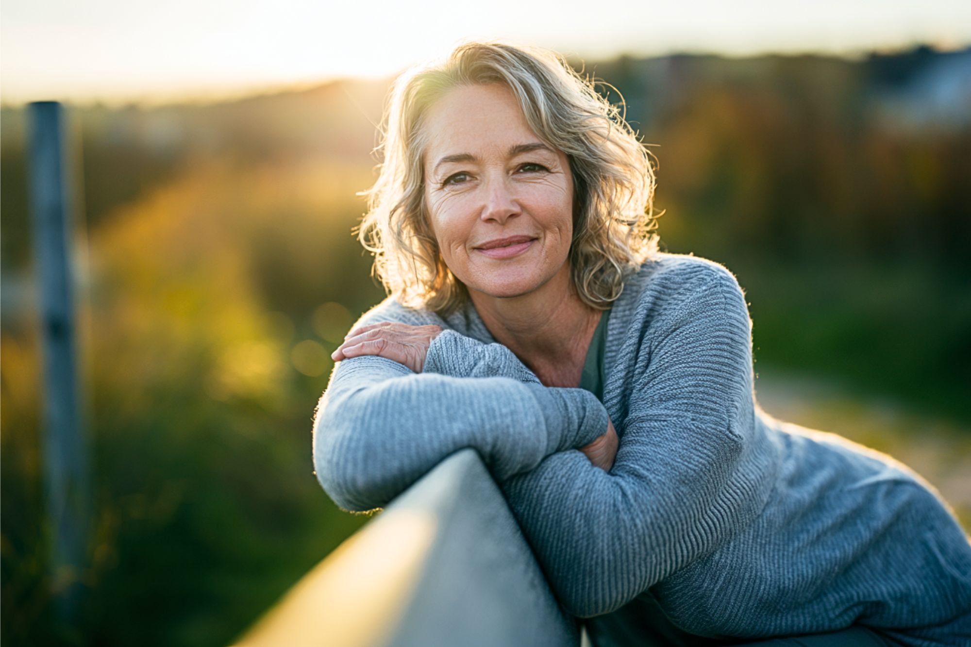 A woman with wavy gray hair and a gray sweater leans on a wooden fence, smiling gently outdoors at sunset with a blurred natural background.