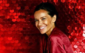 A smiling woman with dark hair pulled back, wearing a red top, stands in front of a vibrant backdrop made of shiny red hexagons.