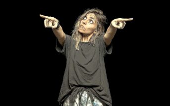 A person with long hair in a loose, dark shirt looks up while pointing both index fingers outward to each side, standing against a plain black background. The image has a cartoon or illustrated effect.