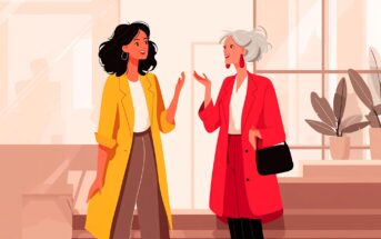 Two women stand indoors, smiling and conversing. One has dark curly hair, a yellow coat, white top, and brown pants; the other has short gray hair, a red coat, white top, black pants, and holds a black bag.