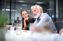 A woman attentively listens to an older man speaking during a meeting in a modern office. Both are seated at a conference table with documents and glasses of water visible.