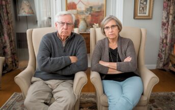 An older man and woman with gray hair sit side by side in beige armchairs, arms crossed and looking serious, in a cozy living room with paintings and decor in the background.