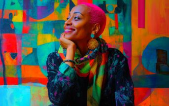 A woman with bright pink hair smiles while looking to the side, resting her chin on her hand. She wears colorful earrings, a multicolored scarf, and sits in front of a vibrant, abstract painted background.