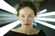 A woman with curly hair and red lipstick stares directly at the camera, standing in front of bright, white lights that radiate outward in a dark, futuristic setting.