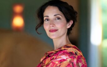 A woman with dark hair and a gentle smile wears a colorful floral-patterned top, standing indoors with soft, warm lighting and a blurred background.