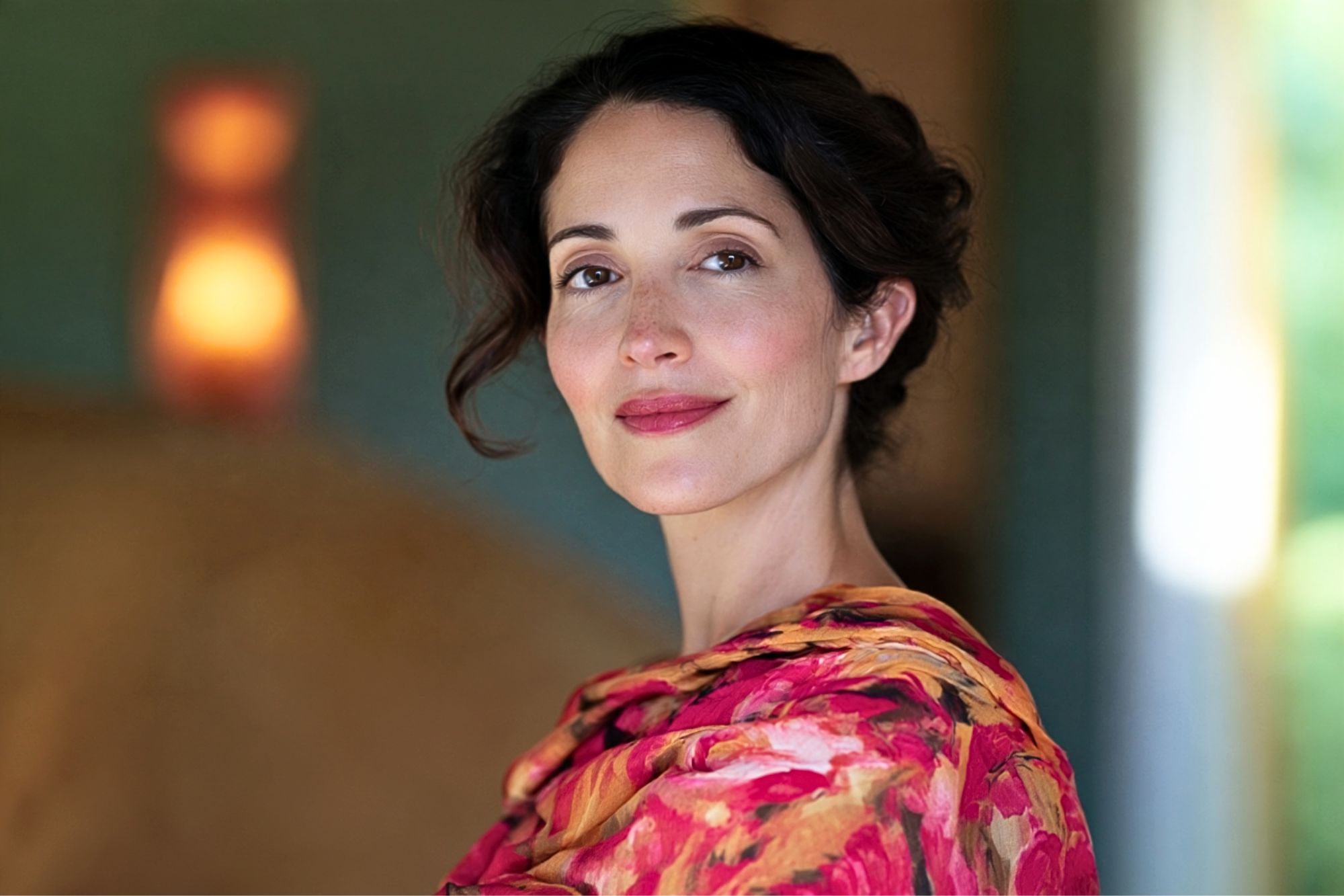 A woman with dark hair and a gentle smile wears a colorful floral-patterned top, standing indoors with soft, warm lighting and a blurred background.