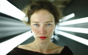 A woman with curly hair and red lipstick stares directly at the camera, standing in front of bright, white lights that radiate outward in a dark, futuristic setting.