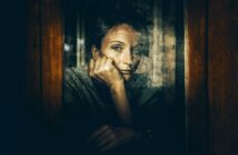 A person with short hair rests their chin on their hand, gazing thoughtfully out of a window. The image is moody and reflective, with muted tones and soft light emphasizing a contemplative mood.