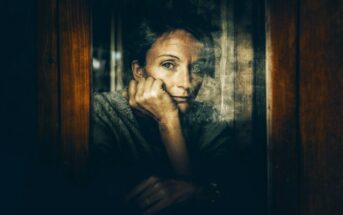A person with short hair rests their chin on their hand, gazing thoughtfully out of a window. The image is moody and reflective, with muted tones and soft light emphasizing a contemplative mood.
