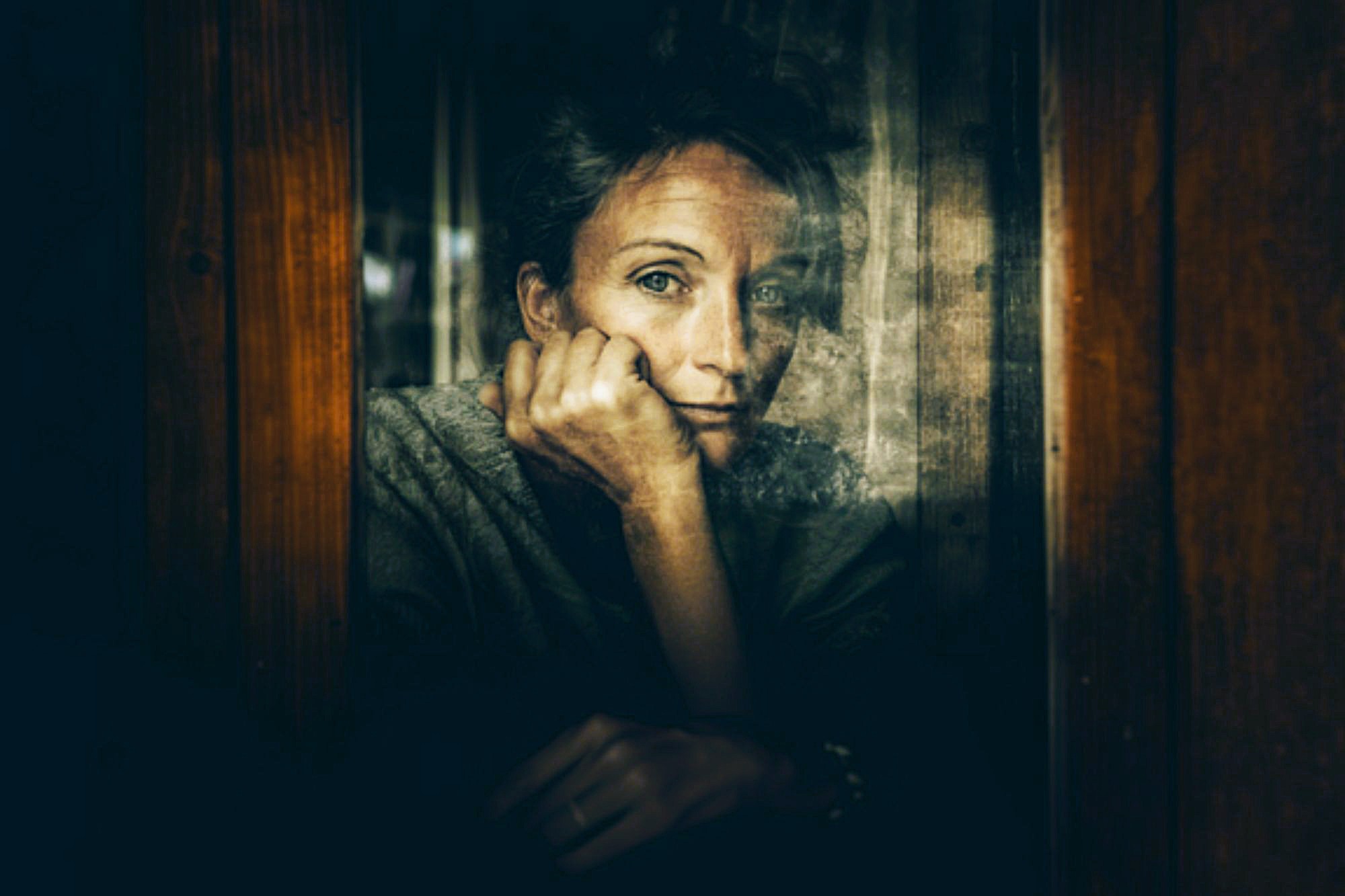A person with short hair rests their chin on their hand, gazing thoughtfully out of a window. The image is moody and reflective, with muted tones and soft light emphasizing a contemplative mood.