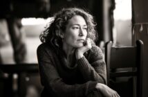 A woman with curly hair rests her chin on her hand, gazing thoughtfully out of a window. She wears a sweater and sits indoors, with soft light illuminating her contemplative expression.