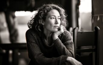 A woman with curly hair rests her chin on her hand, gazing thoughtfully out of a window. She wears a sweater and sits indoors, with soft light illuminating her contemplative expression.