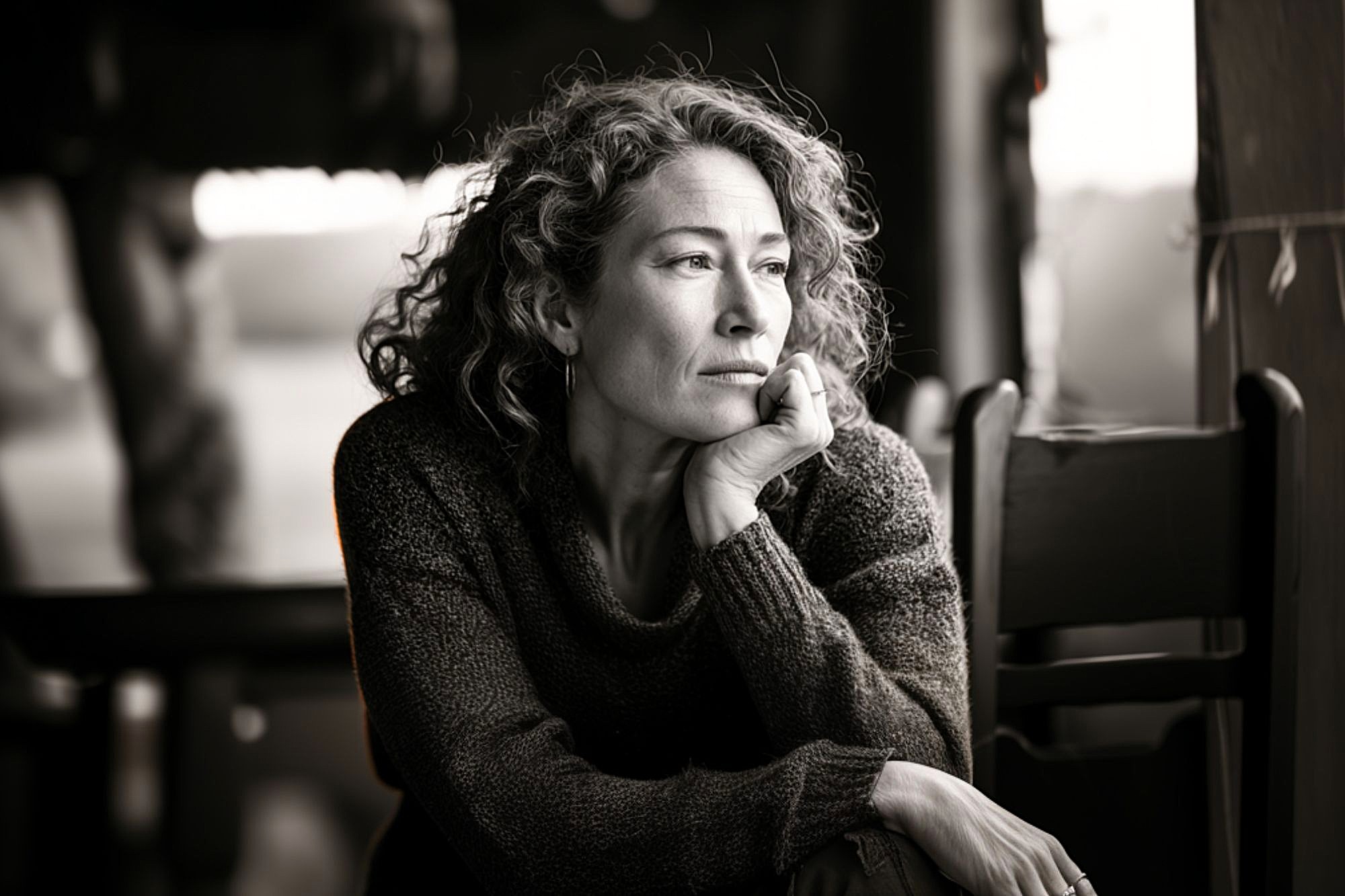 A woman with curly hair rests her chin on her hand, gazing thoughtfully out of a window. She wears a sweater and sits indoors, with soft light illuminating her contemplative expression.