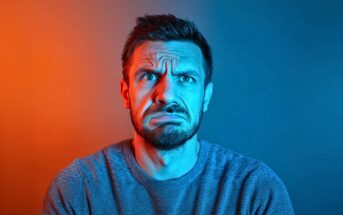A man with a beard and short hair looks confused or concerned, with furrowed brows and a scrunched mouth. Blue and orange lighting split his face, creating a dramatic color contrast.
