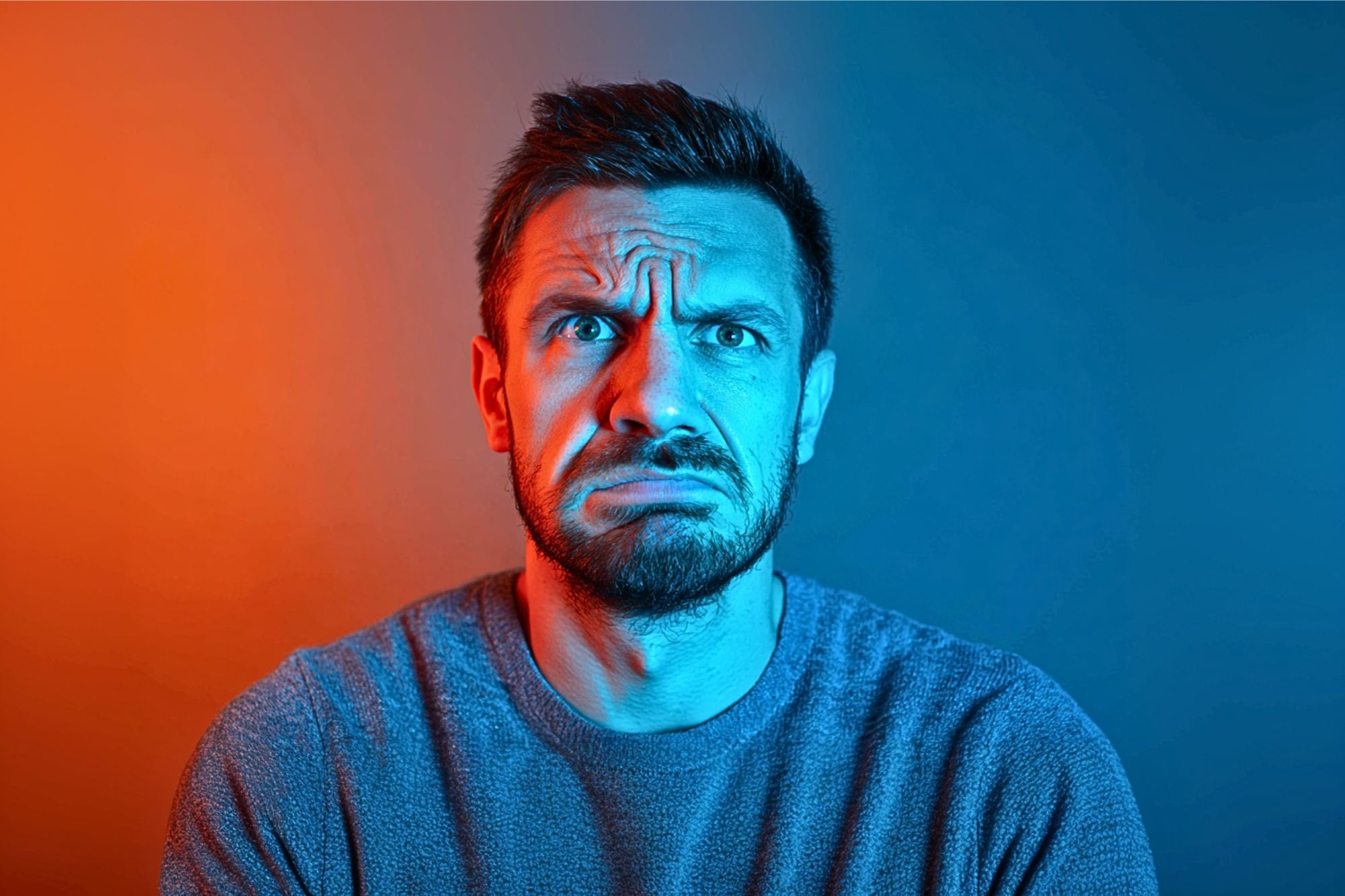 A man with a beard and short hair looks confused or concerned, with furrowed brows and a scrunched mouth. Blue and orange lighting split his face, creating a dramatic color contrast.