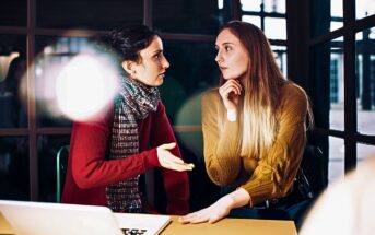 Two women sit at a table in conversation, one gesturing while speaking, the other listening with her hand on her chin. They are indoors, with a laptop on the table and light streaming in through windows.