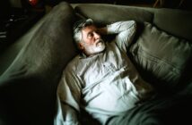 An older man with a gray beard and striped shirt lies on a large gray couch, looking thoughtful and relaxed, with one arm behind his head. The lighting is soft and indirect.