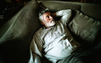 An older man with a gray beard and striped shirt lies on a large gray couch, looking thoughtful and relaxed, with one arm behind his head. The lighting is soft and indirect.