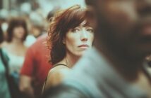 A woman with red hair and fair skin stands in a crowded street, looking directly at the camera with a serious expression. The foreground and background are blurred, highlighting her face.