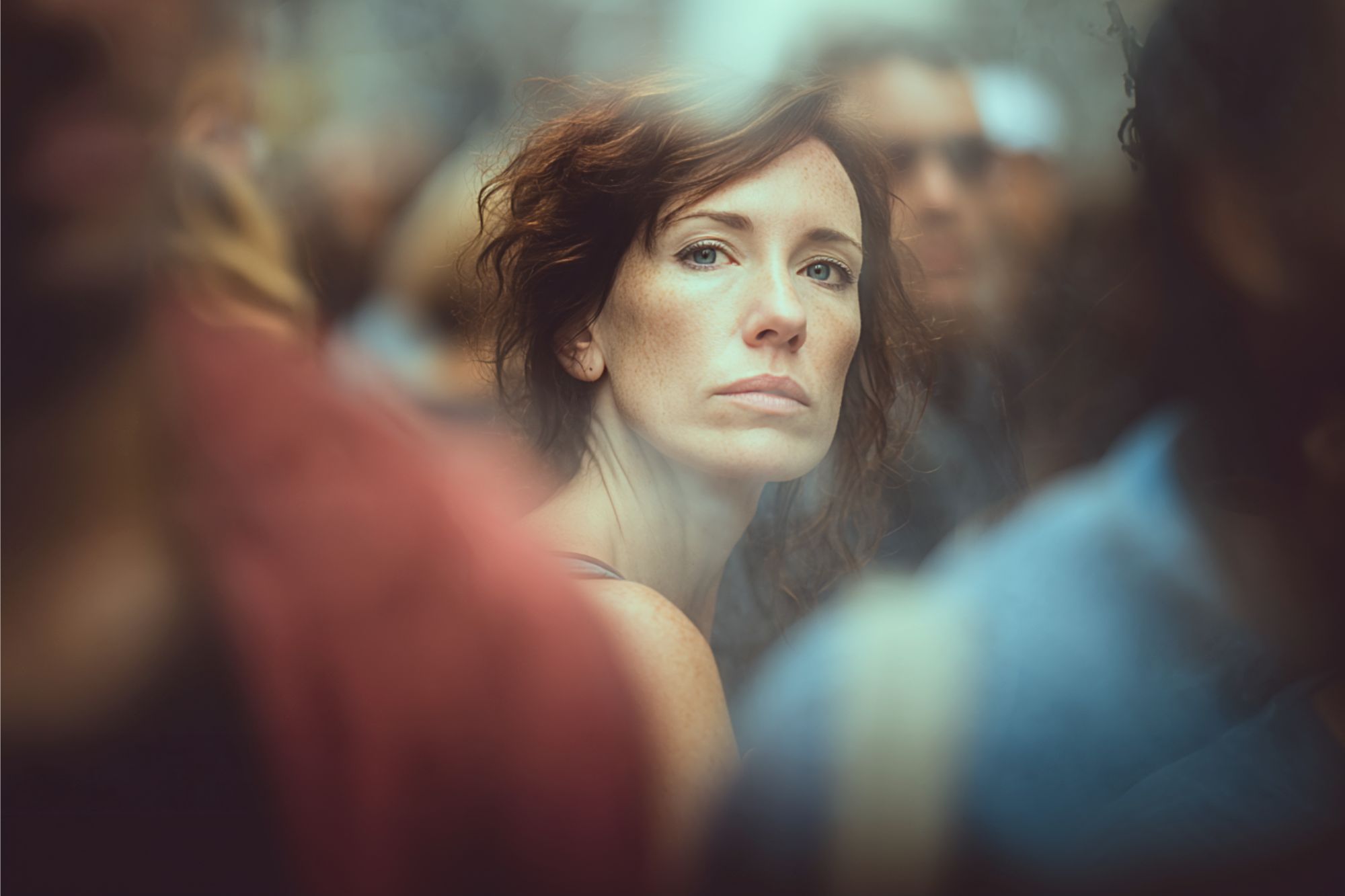 A woman with wavy auburn hair and a serious expression stands among a blurred crowd, looking directly at the camera. The background is out of focus, emphasizing her face.