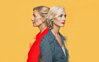 Two women stand back to back against a yellow background. One has gray hair in a bun and wears a red top; the other has short blonde hair and wears a blue polka-dot shirt. Both have red lipstick and look in opposite directions.