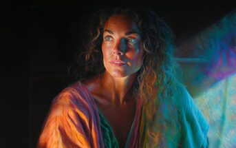 A woman with long, curly hair gazes thoughtfully to the side. She is bathed in dramatic, colorful light that creates a vibrant and moody atmosphere. She wears a draped, multicolored garment against a dark background.