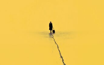 A person in dark clothing stands on a yellow surface, pulling a suitcase along a long crack that runs through the ground, with a minimalist yellow background.