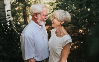 An elderly couple stands close together outdoors, smiling and looking into each other's eyes. They both have white hair, glasses, and are wearing light-colored clothing. Green foliage surrounds them.