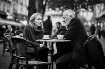 A woman and an older man sit across from each other at an outdoor café table, both with serious expressions, holding coffee cups. People and buildings blur in the background, creating a moody, urban atmosphere.