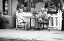 An older man and woman sit across from each other at an outdoor café table, engaged in conversation. The scene is in black and white, with a white picket fence and flowers in the background.