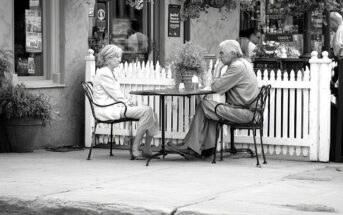 An older man and woman sit across from each other at an outdoor café table, engaged in conversation. The scene is in black and white, with a white picket fence and flowers in the background.