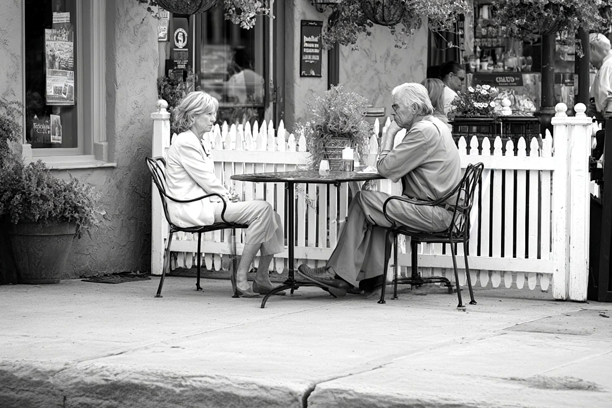 An older man and woman sit across from each other at an outdoor café table, engaged in conversation. The scene is in black and white, with a white picket fence and flowers in the background.