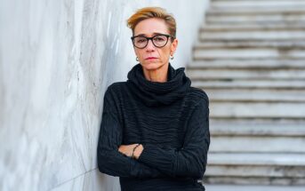 A person with short blond hair and glasses, wearing a black textured top, stands with arms crossed against a white marble wall beside a staircase, looking seriously at the camera.