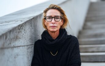 A middle-aged woman with blonde hair, wearing glasses, a black scarf, and a gold necklace, stands outdoors by a concrete wall next to a staircase, looking calmly at the camera.