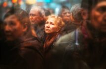 An older woman with light hair and a scarf looks up thoughtfully while standing in a blurred, crowded street scene surrounded by other people.