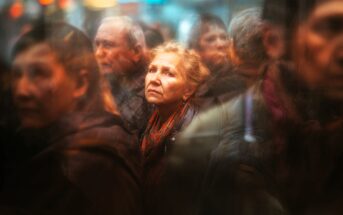 An older woman with light hair and a scarf looks up thoughtfully while standing in a blurred, crowded street scene surrounded by other people.