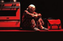 An elderly woman with white hair and glasses sits on the floor in floral pajamas, leaning against a red oven. A red handbag and kitchen pots are beside her, all illuminated by dramatic red lighting.