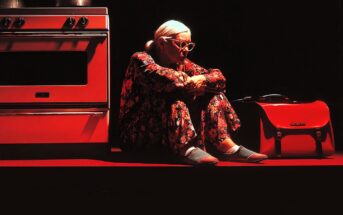 An elderly woman with white hair and glasses sits on the floor in floral pajamas, leaning against a red oven. A red handbag and kitchen pots are beside her, all illuminated by dramatic red lighting.