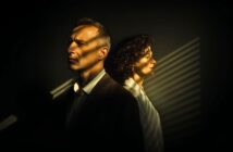 A man and a woman stand back to back in dim lighting, both looking serious. Light from window blinds casts striped shadows across their faces and the dark background.
