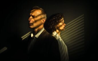 A man and a woman stand back to back in dim lighting, both looking serious. Light from window blinds casts striped shadows across their faces and the dark background.