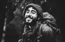 A young man with a beard and curly hair smiles warmly at the camera, wearing a scarf and a backpack. The black-and-white photo gives a feeling of adventure and outdoor exploration.