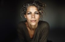A woman with wavy gray hair, light eyes, and a gentle smile looks directly at the camera. She is wearing a loose, dark top and is set against a dark, blurred background.