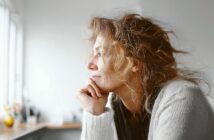 A woman with wavy hair and a light sweater rests her chin on her hand, gazing thoughtfully out of a window in a softly lit room.