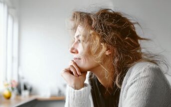 A woman with wavy hair and a light sweater rests her chin on her hand, gazing thoughtfully out of a window in a softly lit room.