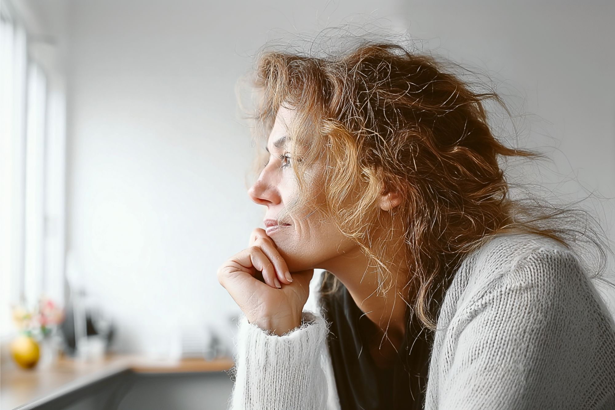 A woman with wavy hair and a light sweater rests her chin on her hand, gazing thoughtfully out of a window in a softly lit room.