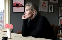 A woman with gray hair, wearing a black sweater, sits at a table by a window, resting her chin on her hand and looking outside. A vase with pink flowers and framed art are visible in the softly lit room.
