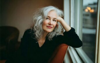 A woman with long gray hair and a black top sits by a window, resting her head on her hand and smiling softly. The background is softly lit and out of focus.